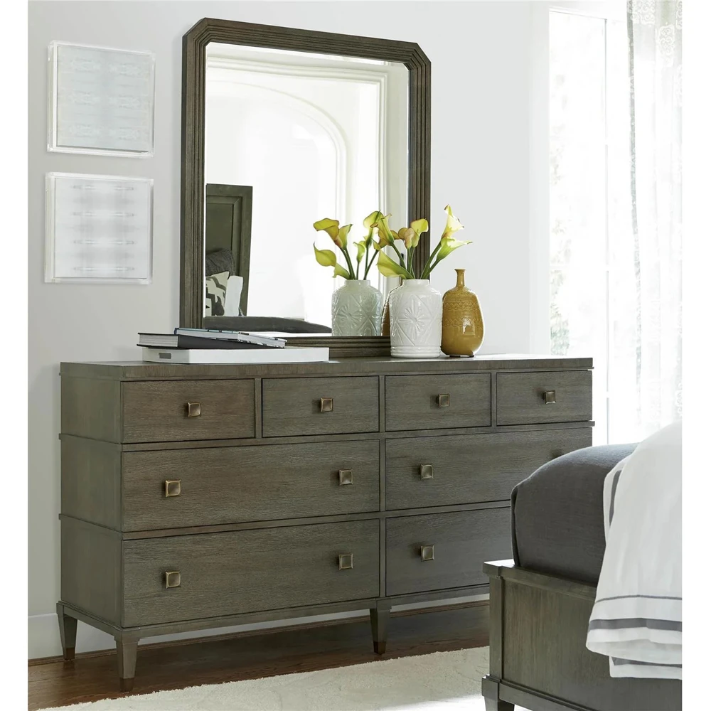 A wooden dresser with nine drawers and gold knobs in a bedroom setting with a mirror and chair.